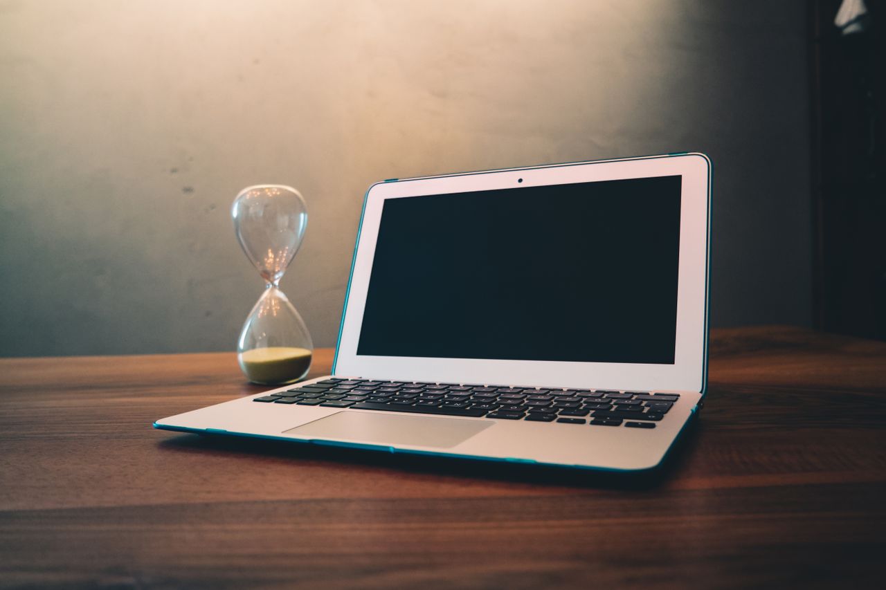 Modern laptop on a wooden desk beside an hourglass, symbolizing time and technology intersection.